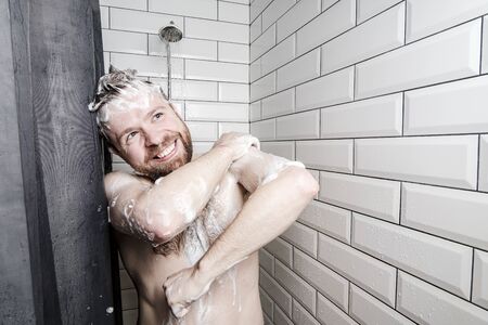 Cute, happy man soaped in foam washes in the shower, under running water, he smiles and looks...
