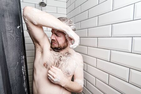 Man in soapy foam, washes in the shower, he sniffs his smelly armpit and is disgusted by the unwashed body. Hygiene concept.の写真素材