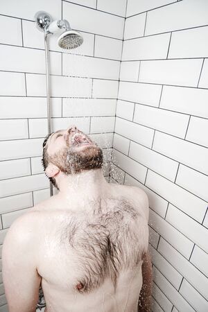 Funny, bearded man stands in the shower with head thrown back and rinses open mouth with warm, running water.の写真素材