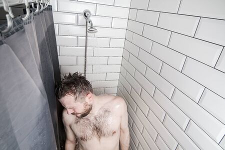 Depressed, frustrated man in the shower, under a stream of warm, running water, looks down and sad.の写真素材