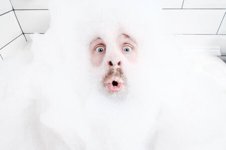 Surprised male face in a lush foam, only eyes and mouth are visible, close-up. Man enjoys lying in the bathtub, with soapy water.の写真素材