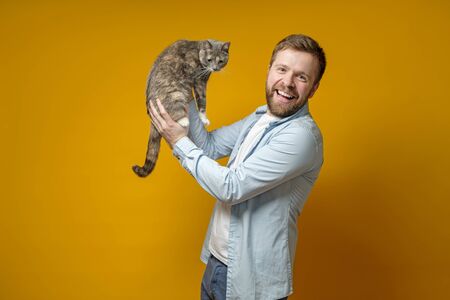 Happy bearded man, owner, holds his miniature cat in hands, looks at the camera and smiles. Concept of love for pets.の写真素材