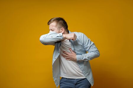 Man in a medical mask coughs in his elbow and holds hand on chest, experiencing pain. Correct sneezing. Concept of the spread of the virus. Isolated on a yellow background.の写真素材