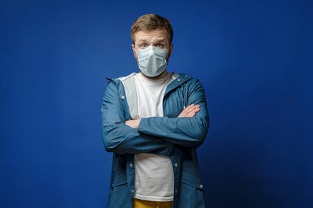 Surprised man in a medical mask stands with arms crossed, on a blue background. Concept of the spread of the virus.の写真素材