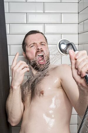 Funny bearded man sings in the bathroom, using a shower head with running water instead of a microphone. Lifestyle.の写真素材