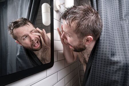 Wet man experiences discomfort, he looks in the mirror and tries to get an eyelash that got into the eye, standing in shower.の写真素材