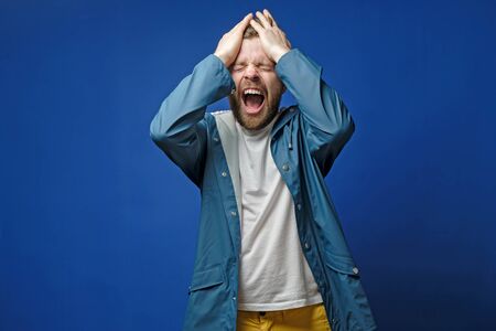 Handsome man is very upset, he holds hands on head and screams loudly, on a blue background. Mental health concept.の写真素材