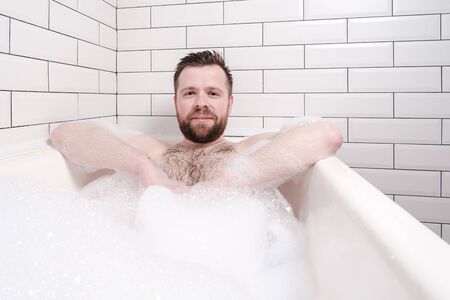 Contented bearded man lies in a bathtub with lush foam and rests.の写真素材