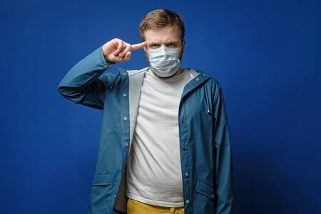 Concentrated man in a medical mask holds finger on temple, trying to find a solution to the problem with the spread of the virus.の写真素材