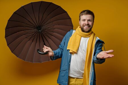 Joyful man in a bright raincoat and knitted scarf stands with an open umbrella, checks the rain with hand, on a yellow background.の写真素材