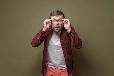 Surprised bearded man, in a burgundy shirt, in amazement holds glasses in hands and looks with big eyes into the camera.の写真素材