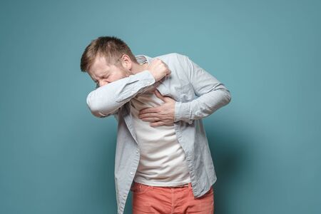Young European man coughs at elbow and holds hand on chest, experiencing pain. Concept of the spread of the virus.の写真素材