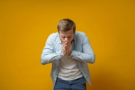 Caucasian man coughs or sneezes while covering mouth with palms. Concept of the spread of the virus. Isolated on a yellow background.の写真素材
