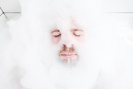 Male face in a lush white foam, only eyes and mouth are visible, close-up. Man relaxes while lying in a bathtub, with soapy water.の写真素材