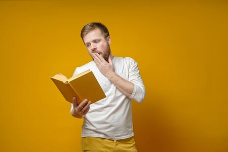 Young, bearded man is fascinated reading a book and emotionally covers his mouth with his hand, on a yellow background.の写真素材