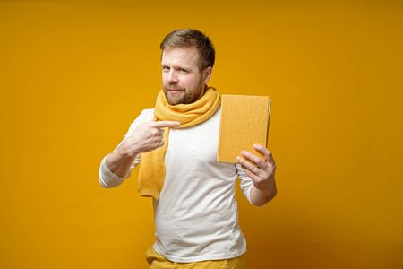 Cute man in a scarf, holds a yellow book in hands, points to it with index finger and looks at the camera.の写真素材