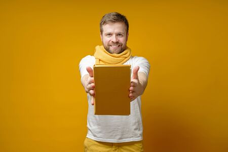Satisfied man in a yellow scarf holds a book on outstretched hands, offers it and smiles at the camera. Education concept.の写真素材