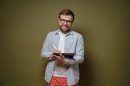 Friendly, bearded man in glasses works with a tablet and stylus pen, smiles and looks at the camera, on a green background.の写真素材