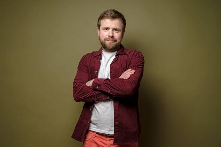 Calm, friendly Caucasian man with a beard stands arms crossed, smiling cute and looking at the camera, on a green background.の写真素材