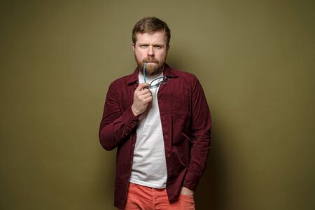 Frowning Caucasian bearded man in a burgundy velvet shirt, looks thoughtfully and sternly, holding glasses near mouth.の写真素材