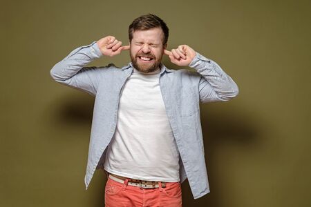 Irritated by a loud sound, the man covers ears with index fingers and grins teeth with displeasure, on a green background.の写真素材
