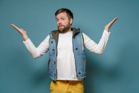 Caucasian bearded man looks guilty, he spreads arms to the sides and apologizes, on a blue...