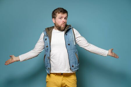 Caucasian man is surprised and bewildered, he throws up hands in perplexity and looks confusedly, on a blue background.の写真素材