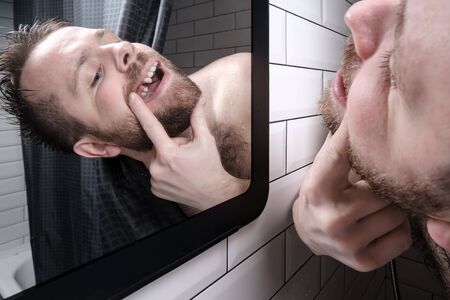 Man opened his mouth and carefully examines the absence of a tooth in mirror, peering out from behind the curtains in the shower.の写真素材