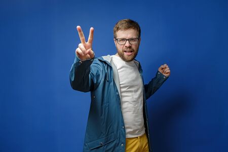Caucasian, bearded man with glasses and a raincoat makes a V-sign with fingers and looks at the camera, on a blue background.の写真素材