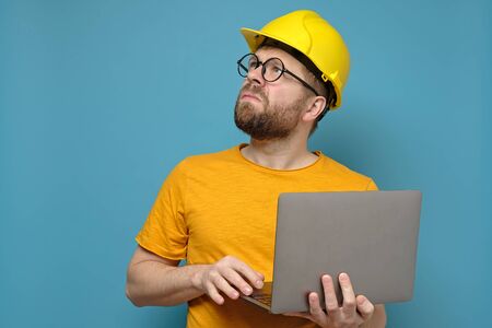 Male foreman or engineer in a construction helmet with a laptop, looking up seriously and displeasedly, on a blue background.の写真素材