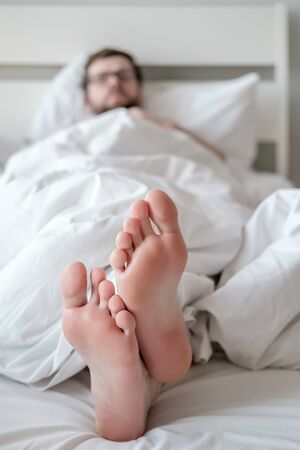 Legs of a sleeping man under a blanket, in bed, in a comfortable apartment, under natural light from the window.