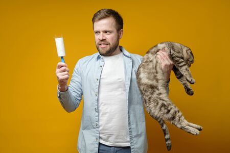 Disgruntled man holds cat in one hand and in the other a roller with adhesive tape in animal hair, which he removed from clothes.の写真素材