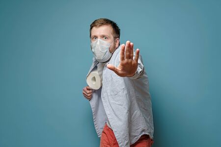 Man in a medical mask hides toilet paper under shirt and makes a stop gesture with hand. Concept of...