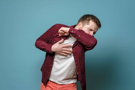 Young, Caucasian man coughs at the elbow and holds hand on chest, experiencing pain. Concept of the spread of a dangerous virus.の写真素材