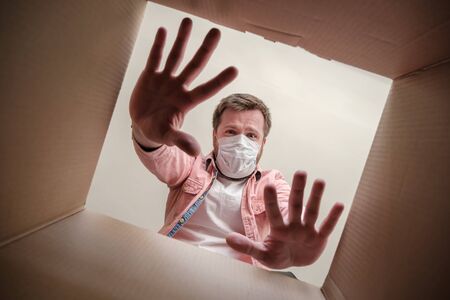 Frightened man in a medical mask unboxing inside view a delivered box, shows a stop gesture with...