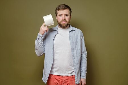 Alarmed man holds in hands a roll of toilet paper, on a green background. Concept of paranoia inflicted with a virus epidemic.の写真素材