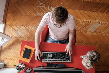 Man works remotely at home on a computer, next to sits his adorable cat. Concept of self-isolation during a pandemic. Lifestyle.の写真素材