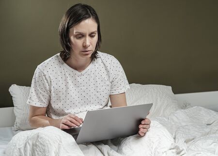 Calm, concentrated woman in pajamas working with a laptop in bed. Concept of self-isolation during a virus epidemic and pandemic.の写真素材