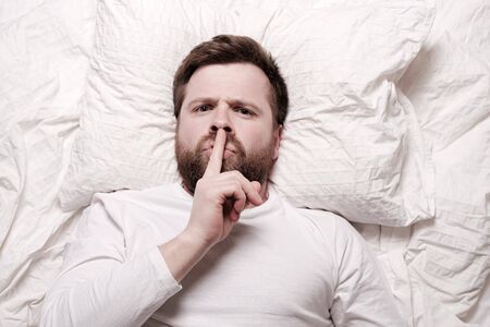Man is lying in bed and puts index finger to lips, expressing silence, not making noise and is staring strictly. Sign language.の写真素材