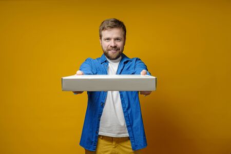 Happy man in a blue shirt came to a meeting with friends with a pizza, he smiles and holds out a box, on a yellow background.の写真素材