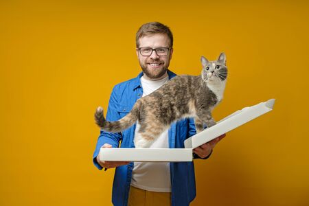 Joyful, smiling man holds in hands an empty pizza box into which his adorable cat climbed, on a yellow background.の写真素材