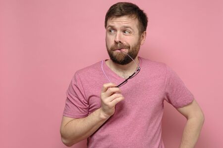 Doubting, bearded man in a pink tee-shirt suspects someone, he holds glasses in mouth and looks oddly in the side.の写真素材