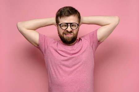 Bearded man with glasses is not in the spirit, he grimaces in exasperation, putting hands behind head, on a pink background.の写真素材