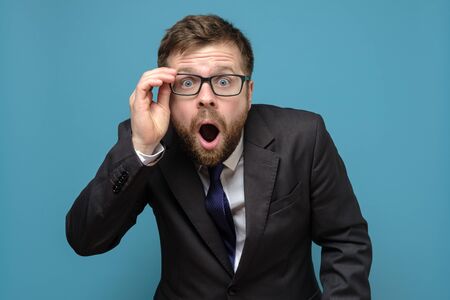 Surprised business man in a suit holds glasses in hand and in amazement opens mouth, looks with big eyes at the camera.の写真素材