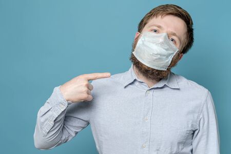 Man points index finger to the medical mask on his face, which is not snug due to the shaggy beard. Quarantine concept.の写真素材