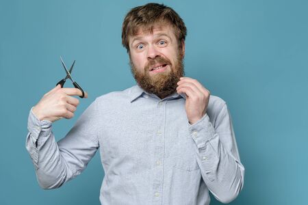 Shaggy man with a beard and scissors in hands, alarmed that he will have to cut his hair independently during quarantine.の写真素材