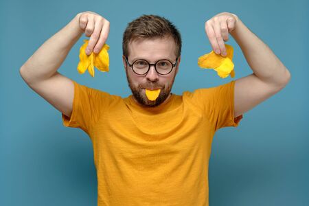 Playful, mischievous man in round glasses and a yellow t-shirt holds dry mango in mouth and hands, on a blue background.の写真素材