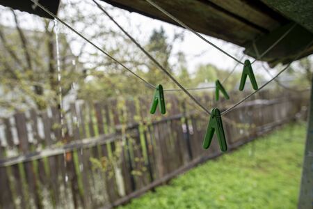 Plastic clothespins hang on a clothesline, under the porch roof, in the rain, against the background of an old fence and garden, on a spring day.の写真素材