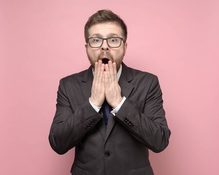 Surprised man with glasses and a suit looks at the camera with big eyes and covers his open mouth with hands.の写真素材