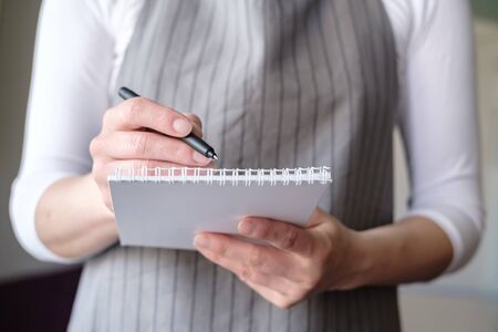 Waitress in a cafe in an apron with a notebook and pen takes an order from the client and writes it in a notepad.の写真素材